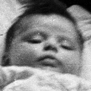 Here's a brief summary of the image: Baby lying in a crib, covered in white fabric.