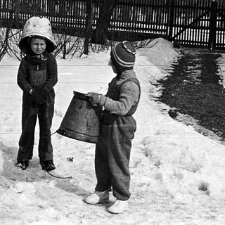 Two children stand in a snowy yard, wearing winter hats.