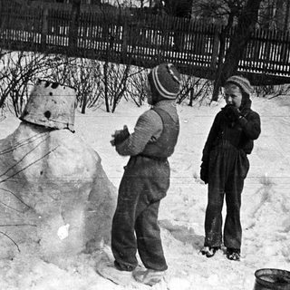 Two children stand near a snowman in a snowy yard.