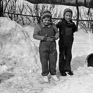 Two children stand in a snowy yard with buckets.