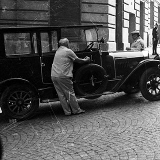 Man assists driver of vintage car on cobblestone street.