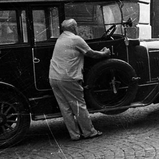 Man assists driver of vintage car on cobblestone street.