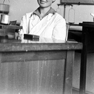 Woman in lab coat stands behind a laboratory table.