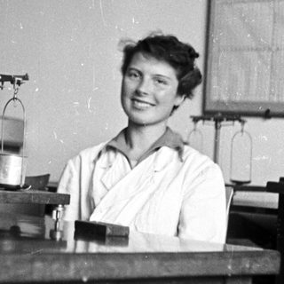 Woman in lab coat smiles behind a laboratory workbench.