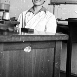 Woman in lab coat smiles behind a laboratory workbench.