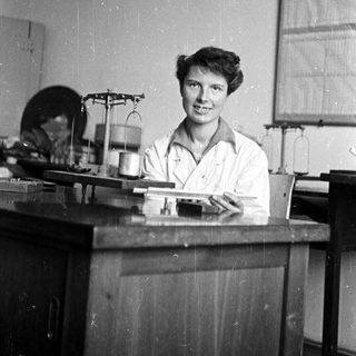 Woman in lab coat smiles behind a workbench.