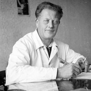 Man in lab coat sits at desk, papers nearby.