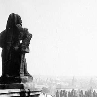 Statue overlooking a city with rooftops and distant buildings.