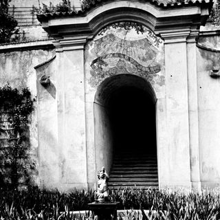 Statue before an arched doorway with stairs and foliage.