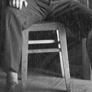 Man in suspenders seated on a wooden stool.