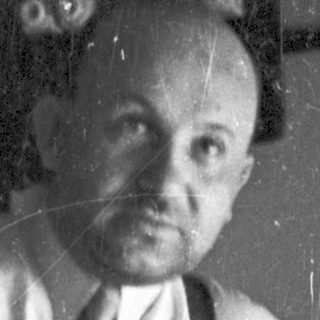 Man in suspenders seated on a wooden stool.