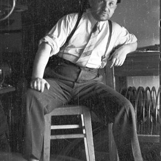 Man in suspenders seated on a wooden stool.