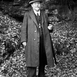 Man in coat stands outdoors near rock face and tree.