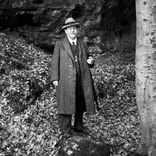 Man in coat stands outdoors near rock face and tree.