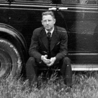 Man sits near vintage car in a wooded area.