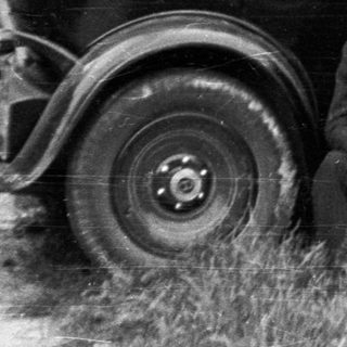 Man sits near vintage car in a wooded area.