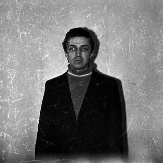 Man in suit stands against textured wall, facing camera.