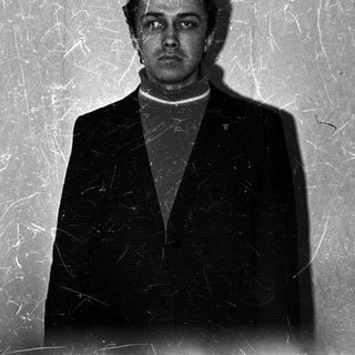 Man in suit stands against textured wall, facing camera.