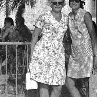 Two women stand near a palm tree, outdoors.