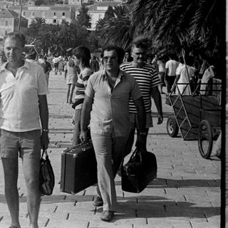 People walking along a harbor with a coastal town backdrop.