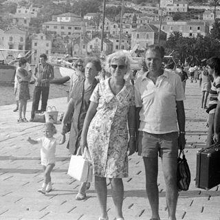 People walking along a harbor with a coastal town backdrop.