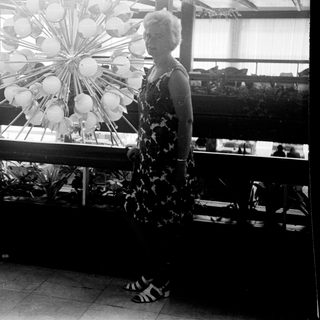 Woman stands near a floral display in a sunlit room.