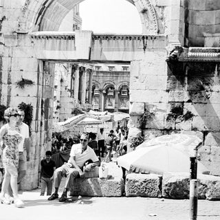 People walk past ancient ruins, a bustling street scene.