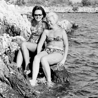 Two women in swimwear sit on rocks by the sea.