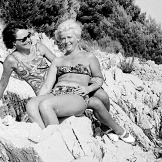 Two women in swimwear sit on a rocky coastline.