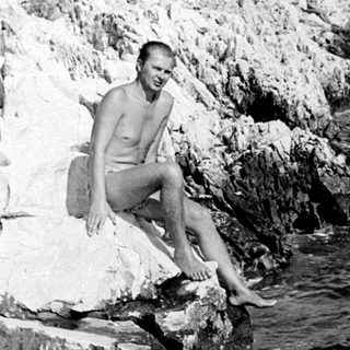 Man sits on rocks by the sea, coastline visible.