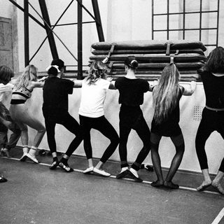 Ballet students stretching in a gymnasium, viewed from behind.