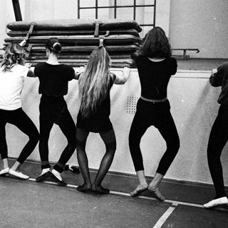 Ballet students stretching in a gymnasium, viewed from behind.
