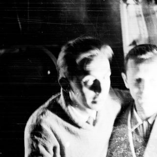 Two men stand near a bookcase in a vintage photo.