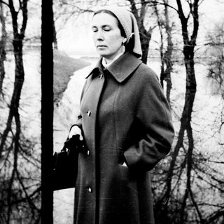 Woman in coat stands on path, another figure in distance.