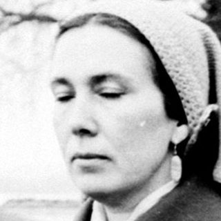 Woman in coat stands on path, another figure in distance.