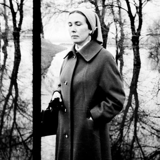 Woman in coat stands on path, another figure in distance.
