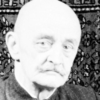 Here's a brief summary of the image: Elderly man in military uniform seated before patterned rug.