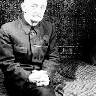 Here's a brief summary of the image: Elderly man in military uniform seated before patterned rug.