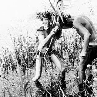 Two shirtless men standing in tall grass near water.
