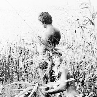 Man stands in tall vegetation near a body of water.
