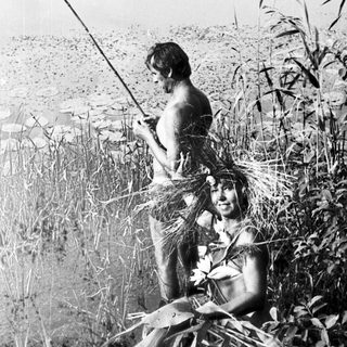 Two people fishing in a lakeside, grassy area.