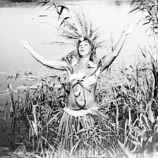 Woman with foliage adornments stands in tall reeds by water.