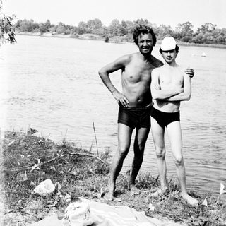 Two men stand by a lake, near a camera.
