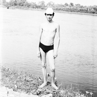 Boy in swimwear stands on riverbank, looking at the camera.