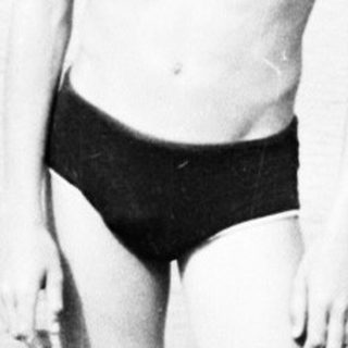 Boy in swimwear stands on riverbank, looking at the camera.
