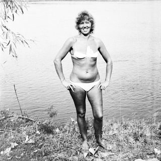 Woman in bikini stands near a lake, smiling.