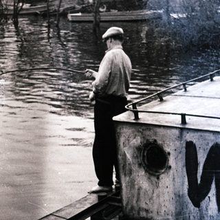 Man stands on a boat near a riverbank.