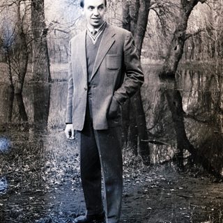 Man in suit stands in a wooded area.