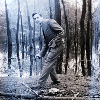 Man in suit crosses a fallen log in a wooded area.