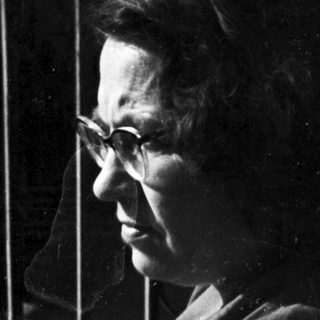 Woman in glasses stands on a balcony overlooking rooftops.